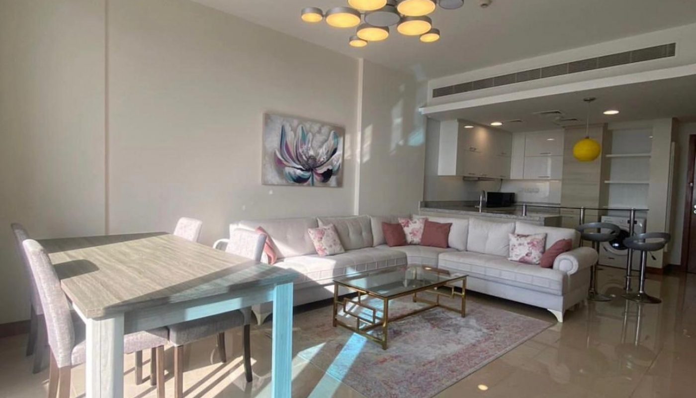 Bright modern living and dining area with a white sectional sofa, glass coffee table, dining table with chairs, and an open kitchen in the background.