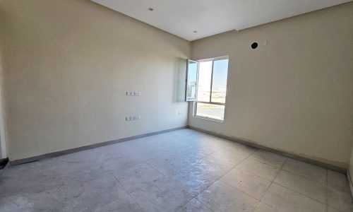 Luxurious villa for sale: Empty room with beige walls, gray tiled floor, and a large window letting in natural light. Located in Hamala.