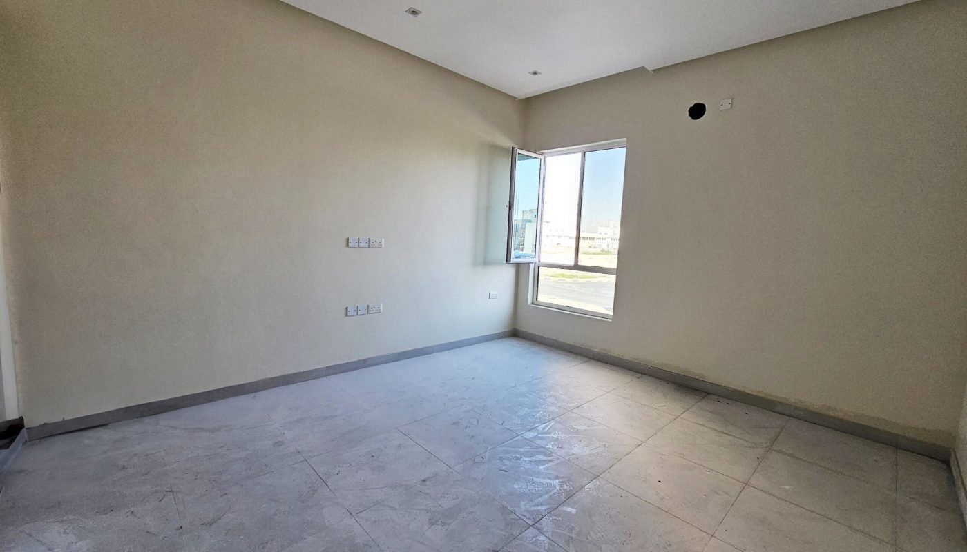 Luxurious villa for sale: Empty room with beige walls, gray tiled floor, and a large window letting in natural light. Located in Hamala.