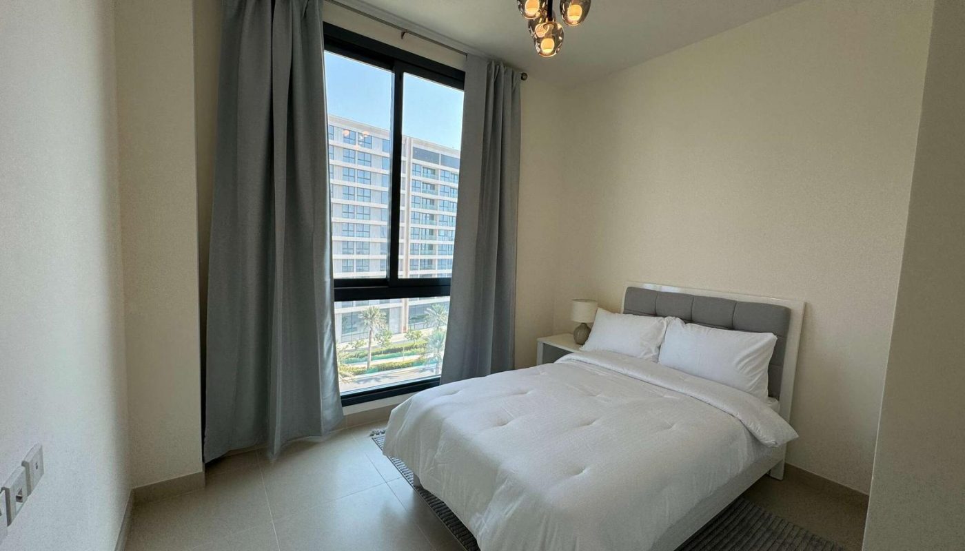 Minimalist bedroom with a double bed, gray headboard, white bedding, bedside table, lamp, large window with gray curtains, and a modern ceiling light fixture.