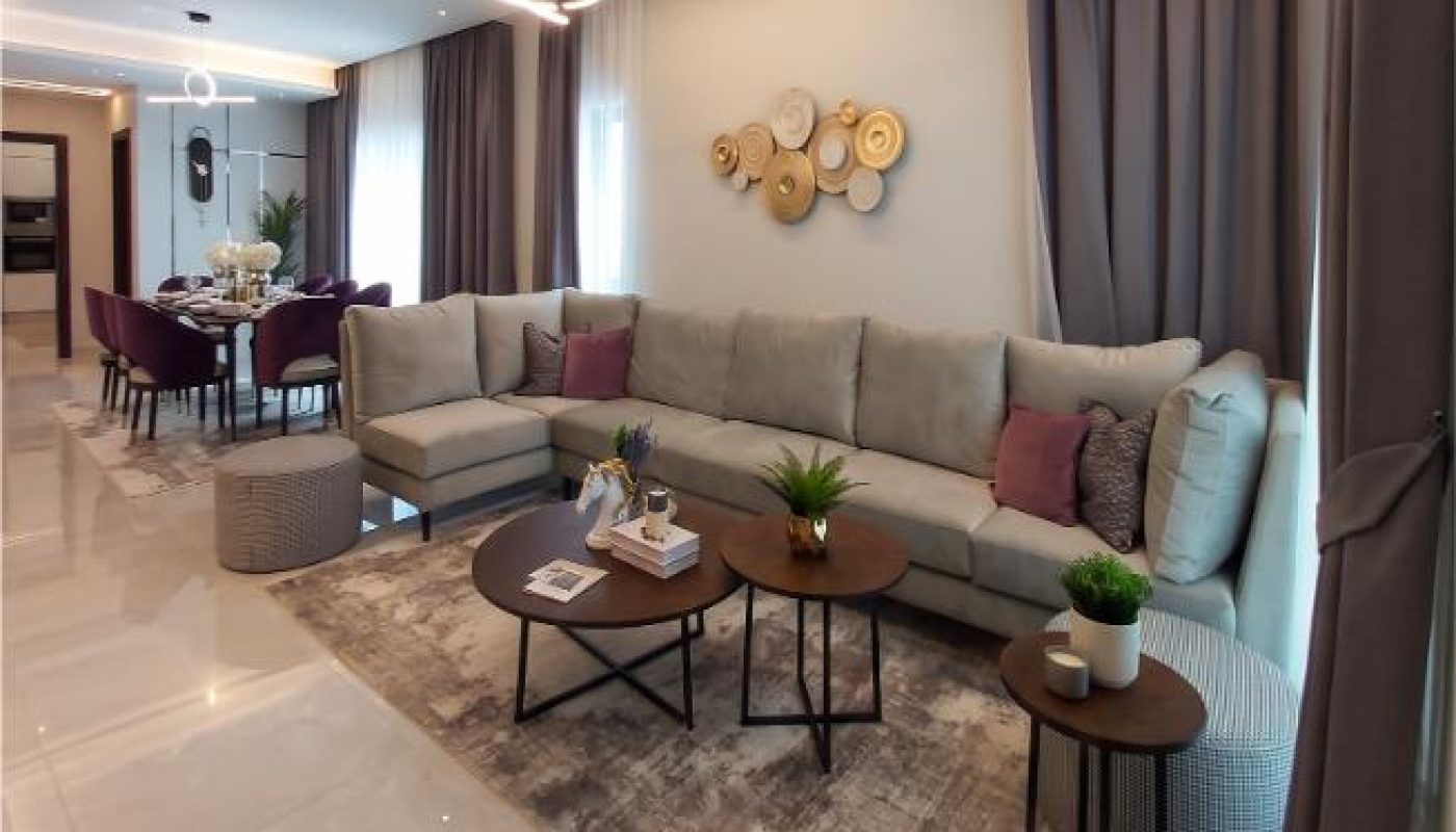 Modern living room with a large L-shaped sofa, round coffee tables, decorative wall art, plants, and floor-to-ceiling curtains; dining area visible in the background.