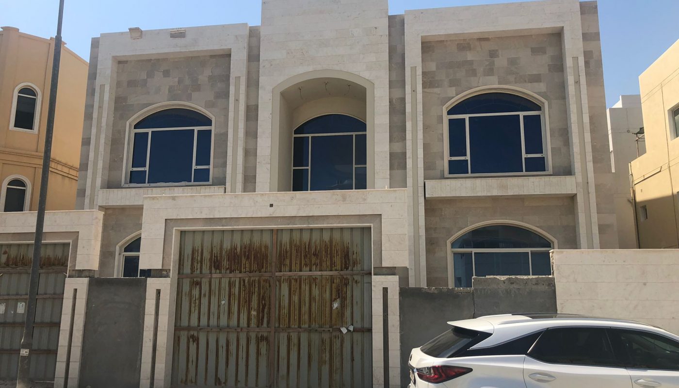 A modern two-story building with large arched windows, a rusted metal gate, and a white SUV parked in front on a sandy street.