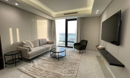 A modern living room in this AMWAJ flat for sale features a beige sofa, a black armchair, two round coffee tables, and a wall-mounted TV, with a large window offering a view of the sea.