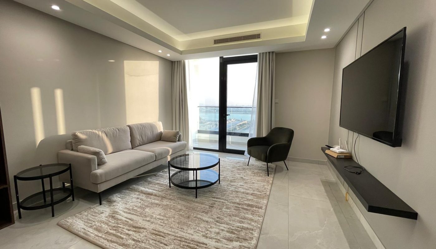 Modern living room with a beige sofa, black armchair, two round glass tables, wall-mounted TV, large window with a sea view, and a light patterned rug on a tiled floor.