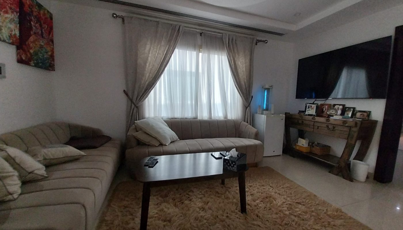 A modern living room with beige sofas, a coffee table, a TV on the wall, framed photos on a wooden console, and a window with curtains letting in natural light.