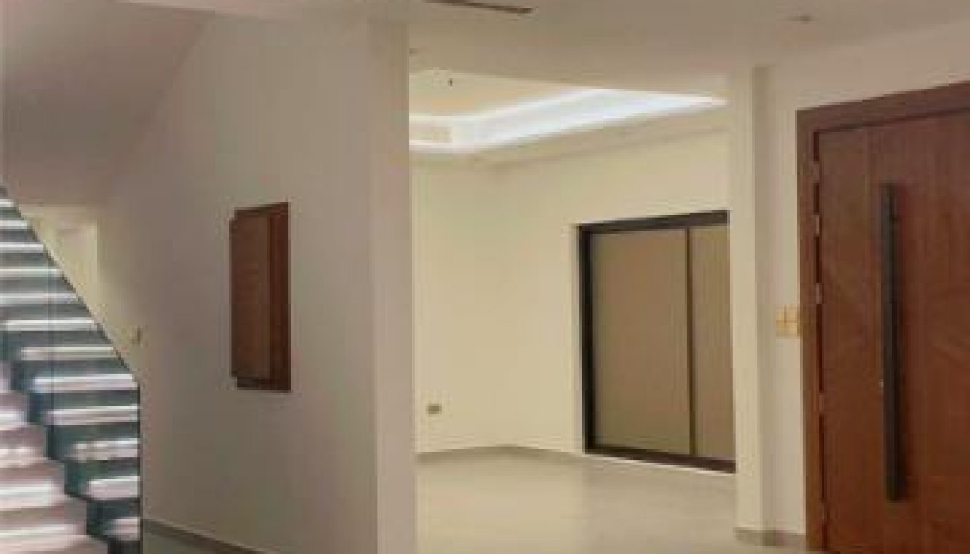 Modern empty interior with light-colored tile flooring, a wooden front door, and a staircase with illuminated steps on the left. Walls and ceiling are white, with recessed lighting throughout.