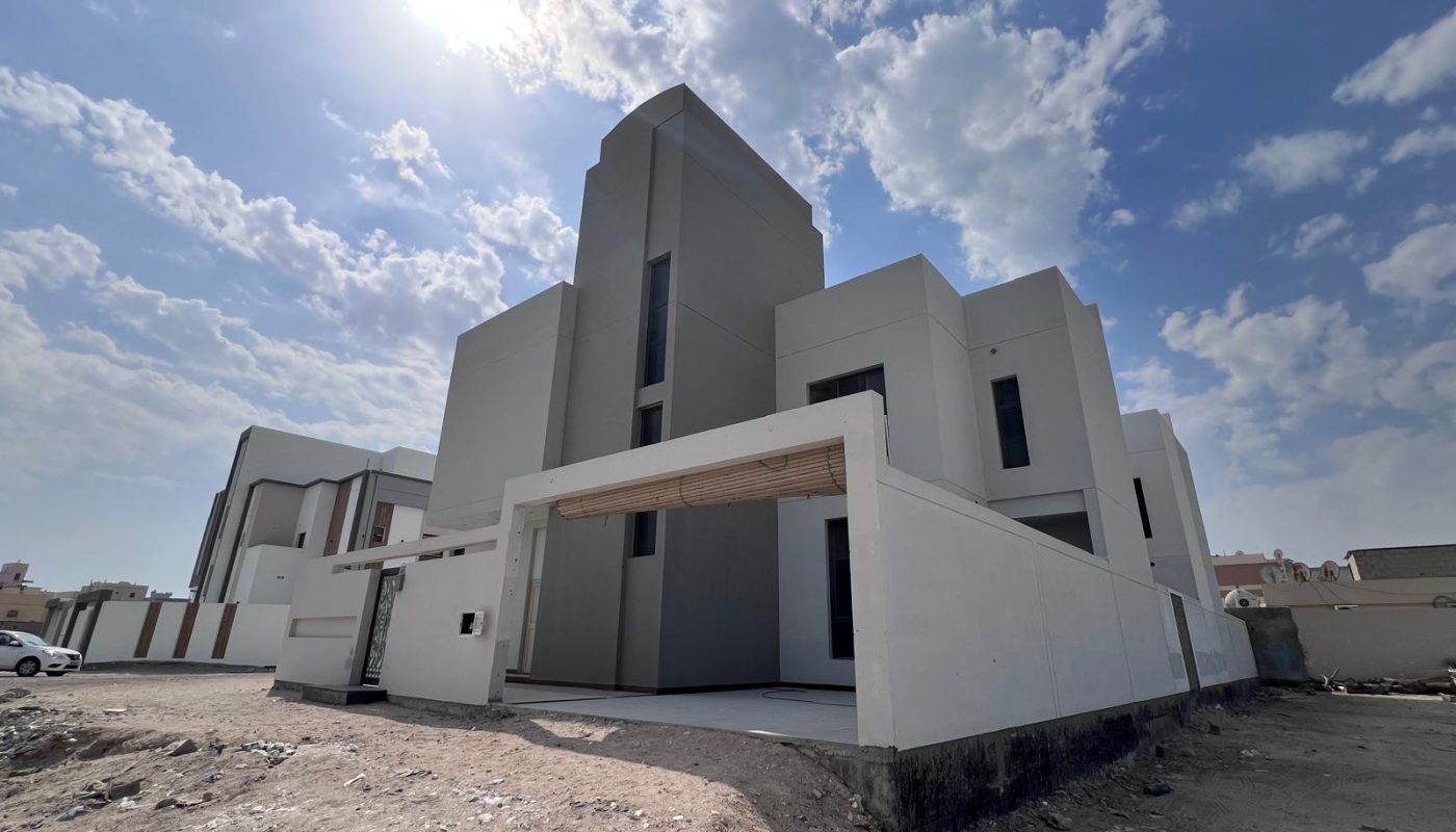 A modern, multi-story white building with clean geometric lines under a partly cloudy sky, surrounded by bare ground and neighboring structures.