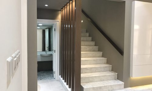 Modern interior featuring a white marble staircase with wooden slats, leading up to the next floor in this luxury villa. A bathroom can be seen at the end of a hallway to the left of the stairs in this stunning Janabiya home that boasts 3 bedrooms.