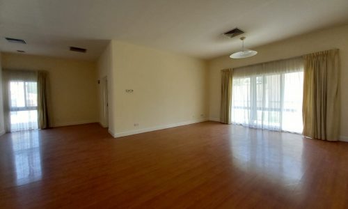 A spacious, empty room with wooden flooring, large windows covered by drapes, and plain white walls.