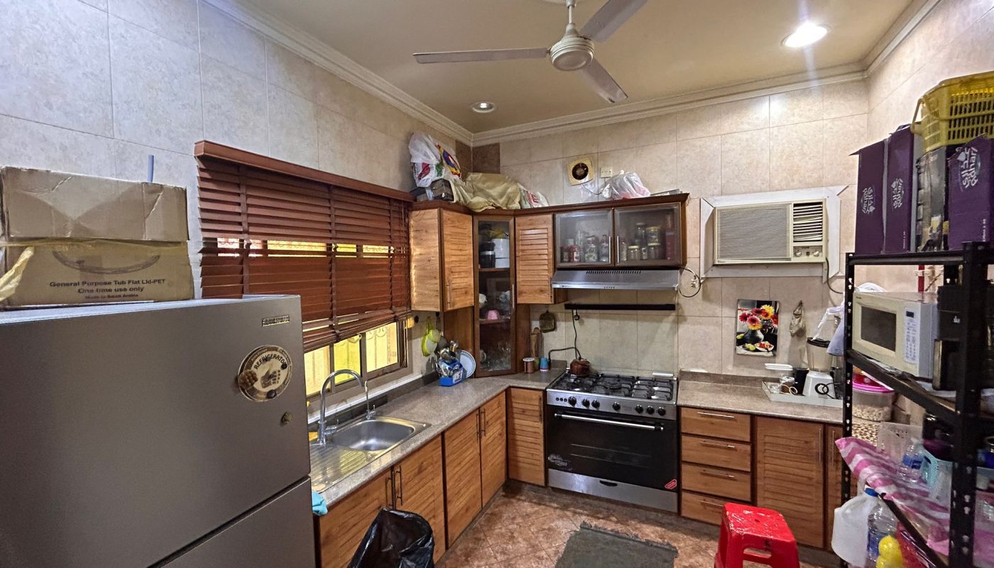 A compact kitchen with wooden cabinets, a refrigerator, stove, sink, wall-mounted air conditioner, shelves, and a red plastic stool.