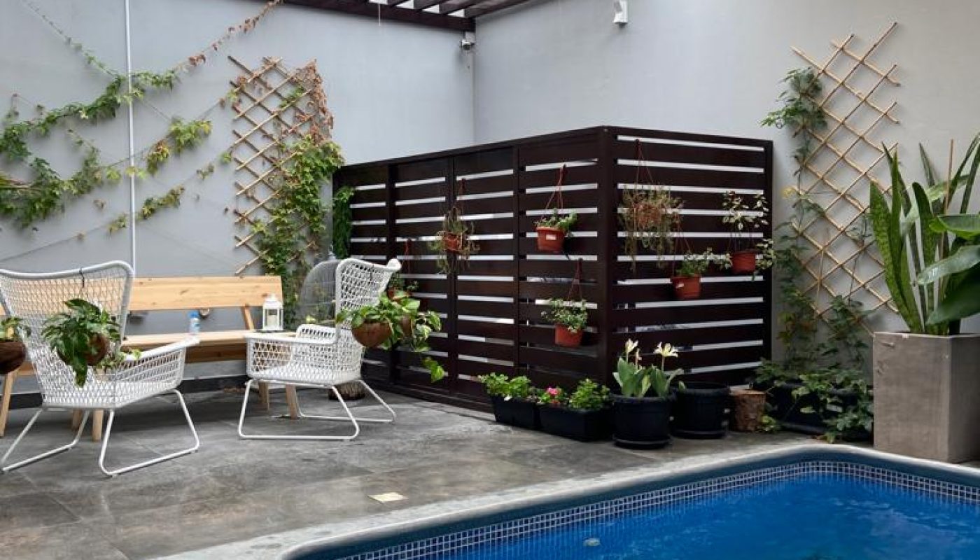 Outdoor patio with a small pool, modern seating, potted plants on a wooden privacy screen, and shade sails above.