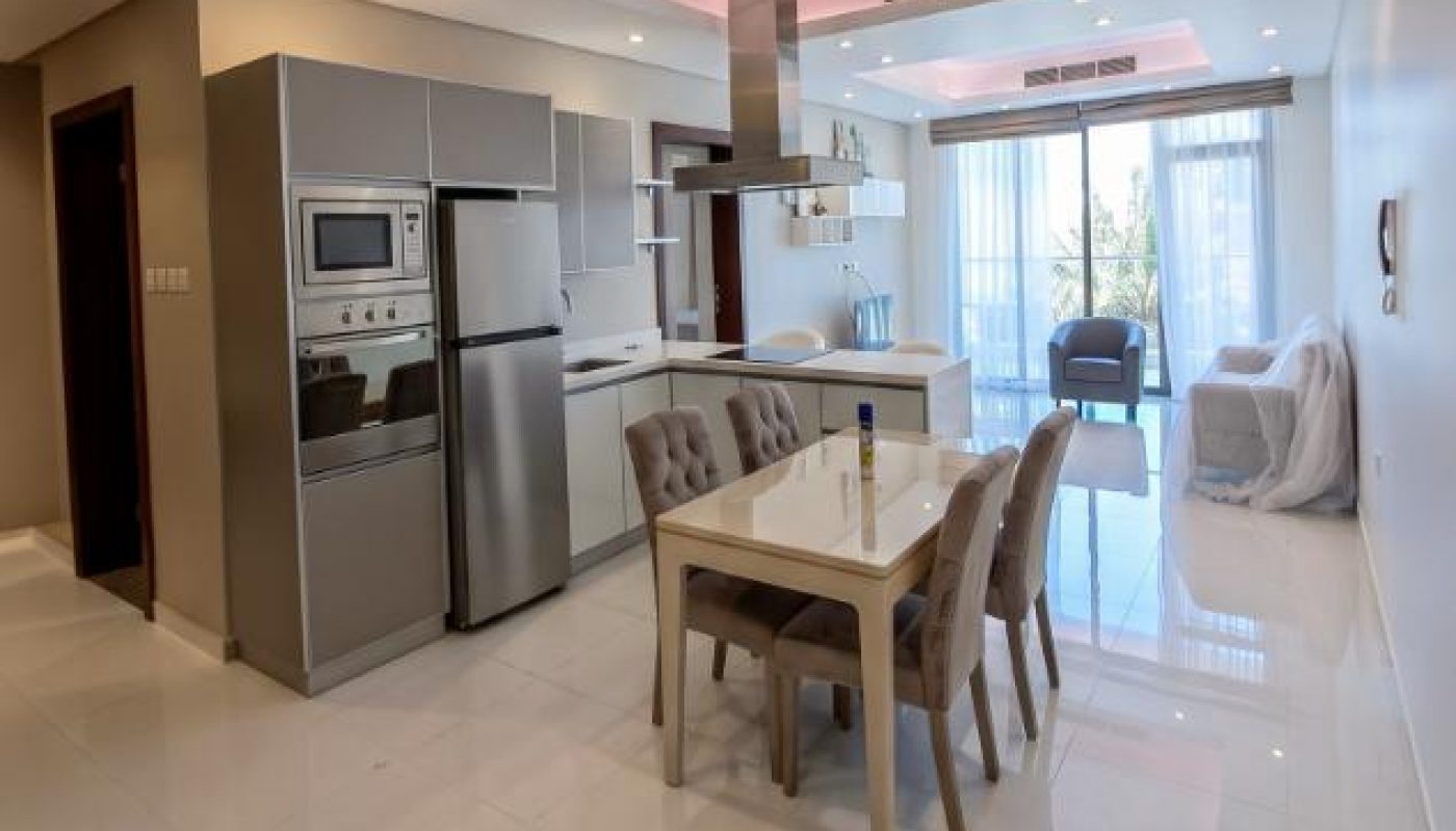 Modern kitchen and dining area with stainless steel appliances, a rectangular table with four chairs, glossy white floors, and large windows letting in natural light.