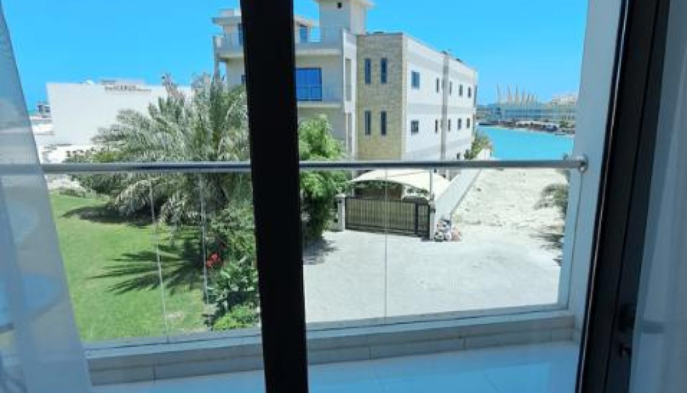 View from a balcony through glass doors, showing a garden, a modern building, a gate, and water with boats in the background under a clear blue sky.
