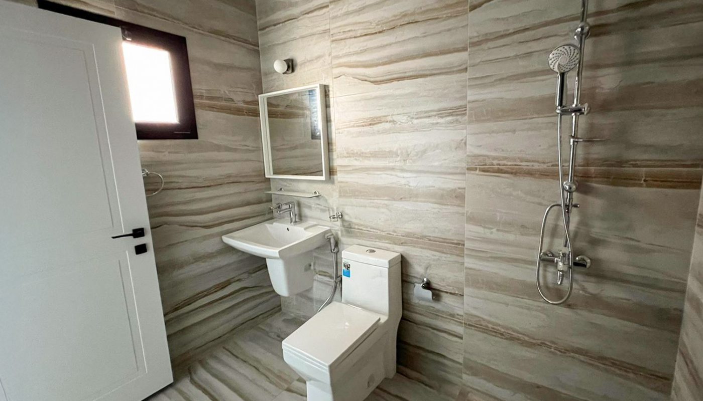 Modern bathroom with beige marble-patterned tiles, a toilet, wall-mounted sink with mirror, shower fixture, and a small window near a white door.