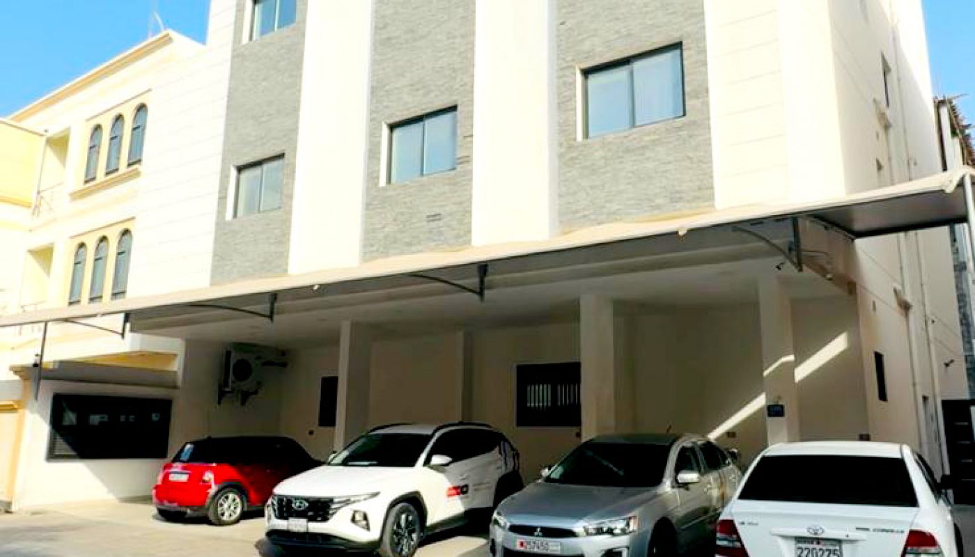 Three-story modern apartment building with four cars parked in front under a carport, photographed on a clear, sunny day.