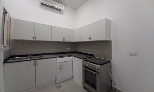 A small, modern kitchen in a luxury villa with white cabinets, black countertops, a stainless steel sink, a dishwasher, a gas stove, and white walls. An air conditioning unit is mounted above the cabinets. This elegant space complements the villa's 3 bedrooms located in Janabiya.