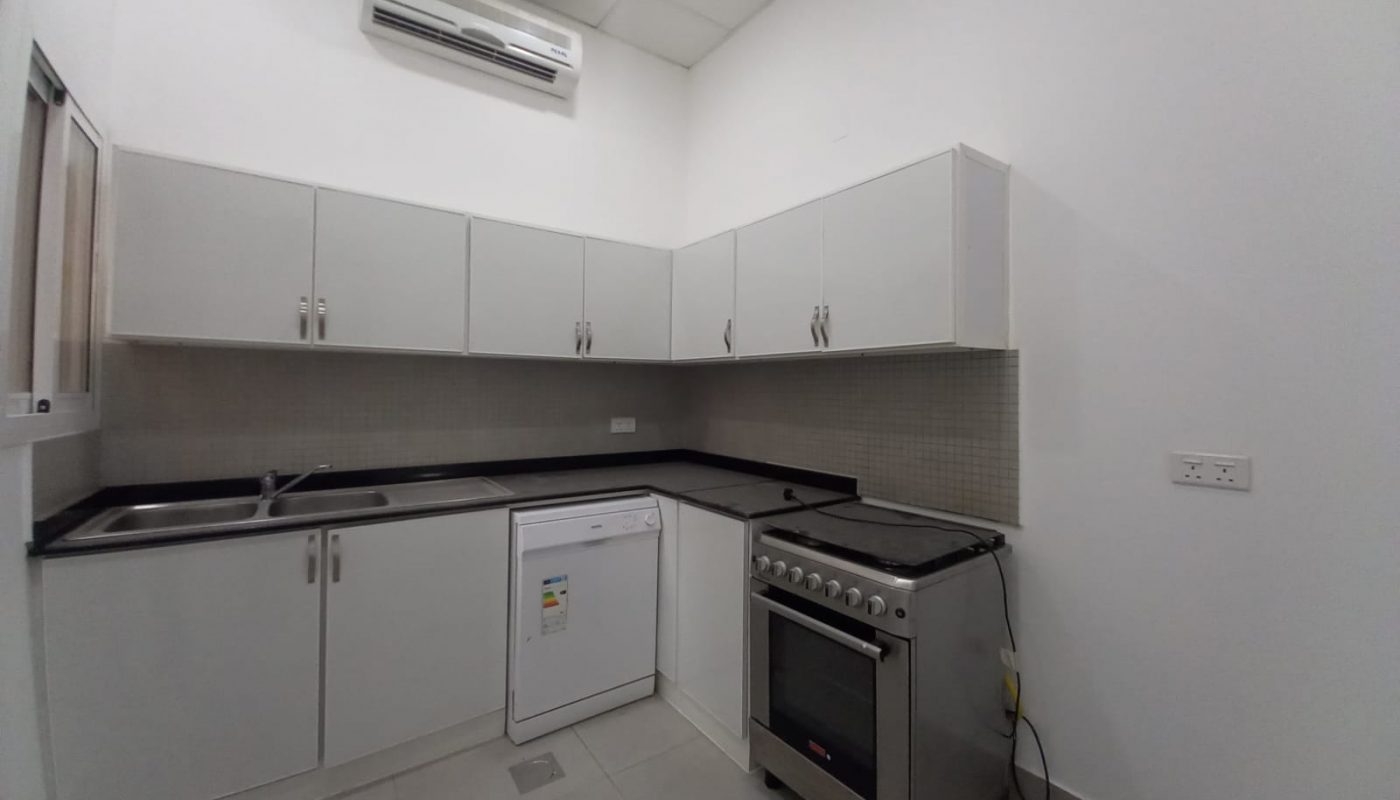 A small, modern kitchen in a luxury villa with white cabinets, black countertops, a stainless steel sink, a dishwasher, a gas stove, and white walls. An air conditioning unit is mounted above the cabinets. This elegant space complements the villa's 3 bedrooms located in Janabiya.