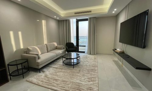 A modern living room in this AMWAJ flat for sale features a beige sofa, a black armchair, and two round coffee tables on a light rug. A flat-screen TV is mounted on the wall, and a large window offers a stunning view outside.