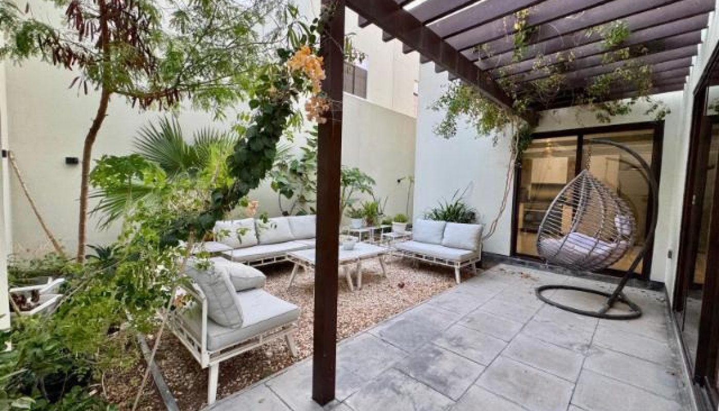 Outdoor patio with a pergola, white cushioned seating, plants, a hanging egg chair, and a mix of gravel and paved flooring.