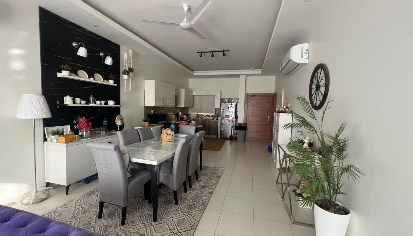 Modern open-plan living and dining area with a glass dining table, gray chairs, white kitchen cabinets, decorative shelves, indoor plants, and a wall clock.