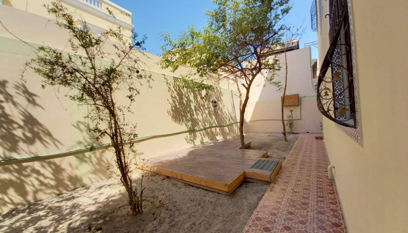 A small courtyard with a tiled walkway, a tree, and a wooden platform surrounded by beige walls under a clear blue sky.
