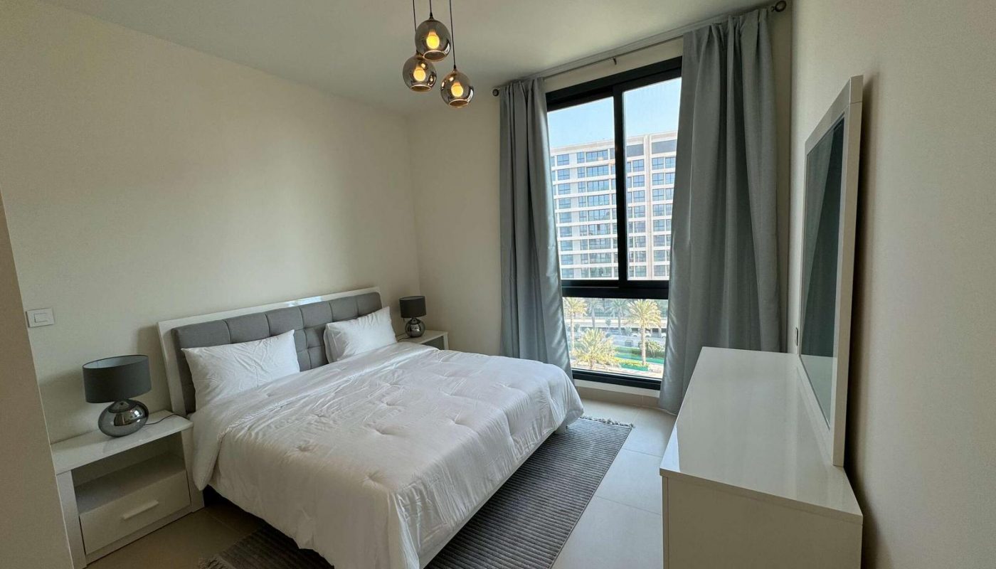 Modern bedroom with a neatly made bed, bedside tables, a dresser, and a large window with gray curtains overlooking tall buildings.