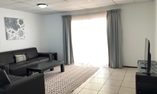 A minimalistic living room with a black sofa set, a dark coffee table, a patterned rug, a flat-screen TV, and a large window with grey curtains. The room has white walls and a tiled floor.