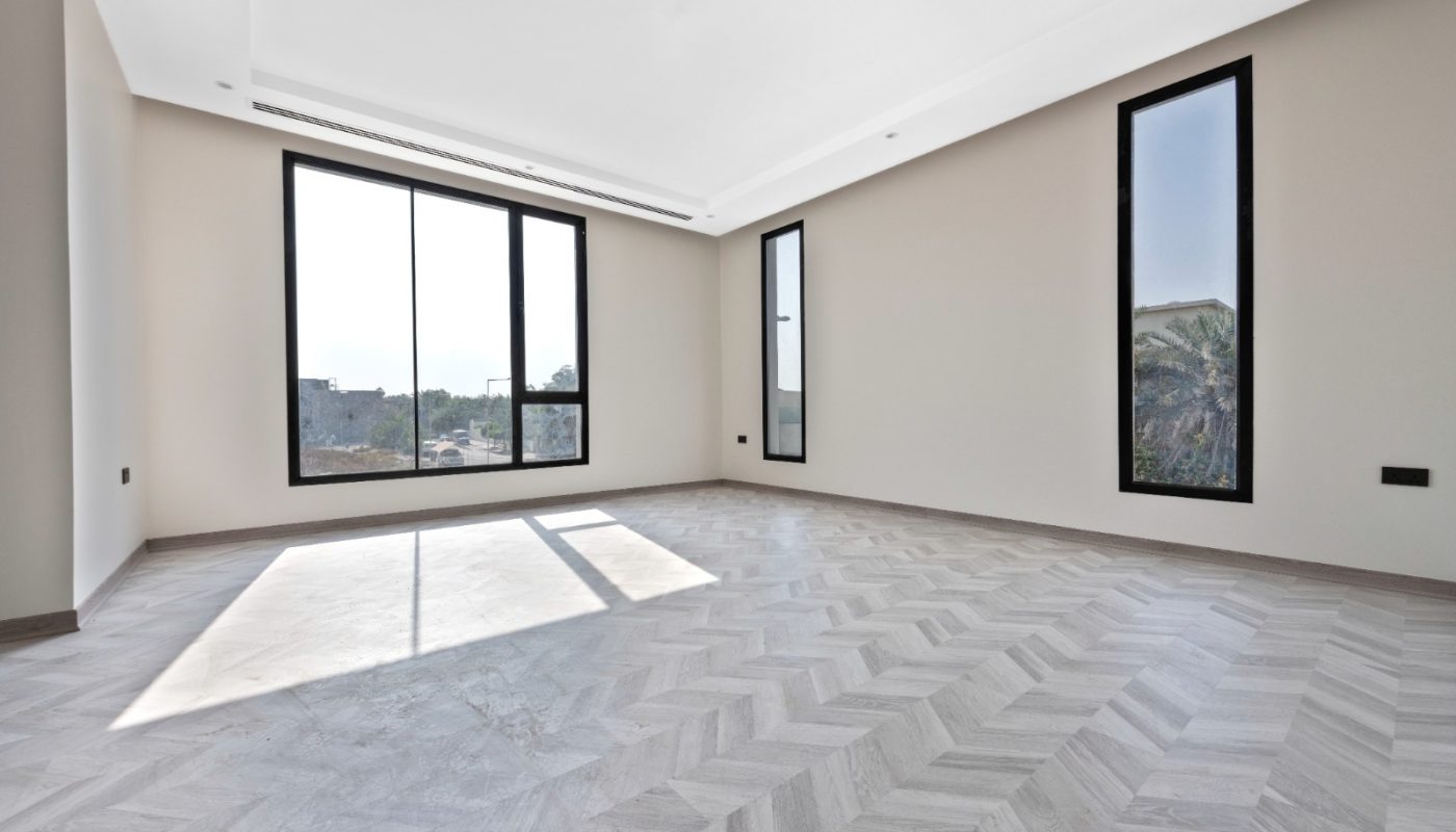 Empty modern room with light-colored chevron-patterned flooring, beige walls, a large window, and two tall narrow windows letting in natural light.