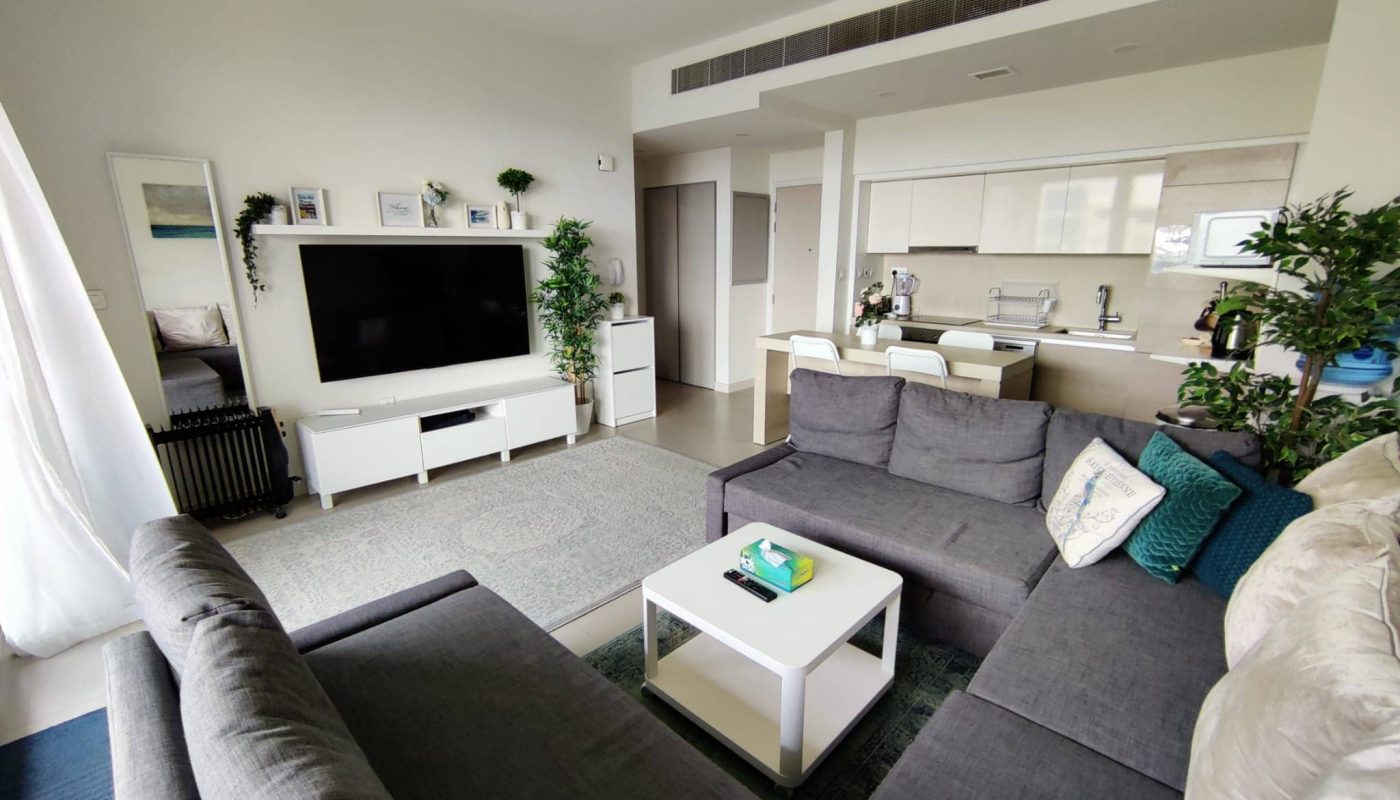 Modern living room with a large gray sectional sofa, white coffee table, flat-screen TV, and open kitchen area in the background. Green plants and neutral decor are visible throughout the space.
