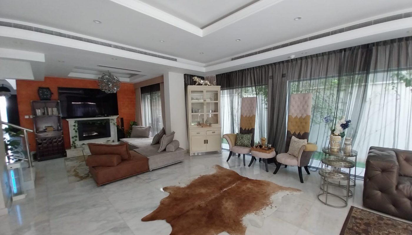 Modern living room with marble floors, a brown rug, large windows with sheer curtains, contemporary furniture, and a wall-mounted TV above a fireplace.