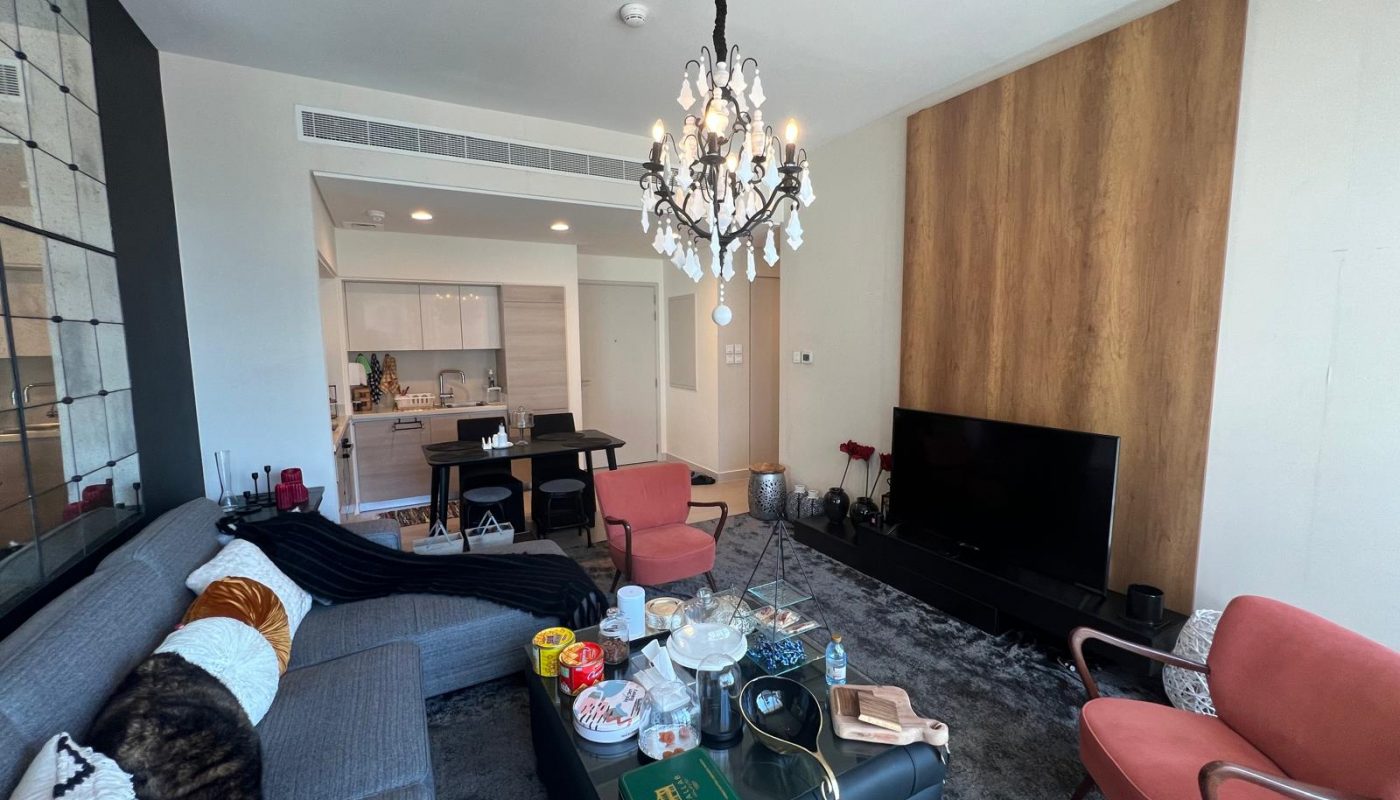 A modern living room with a large gray sofa, black coffee table with various items, TV on a stand, chandelier, and an open kitchen in the background.