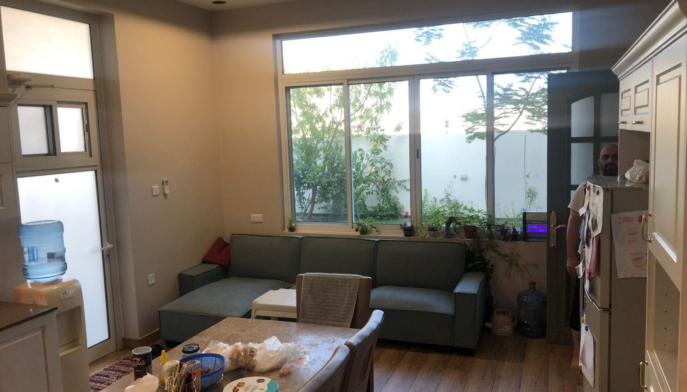 A living room with a large window, sofa, table with assorted items, water dispenser, and kitchen area; greenery is visible outside the window.