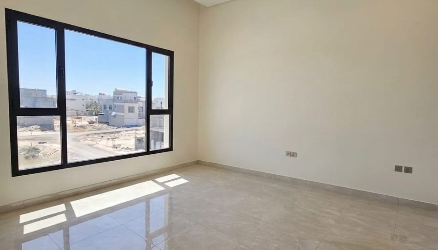 Empty, unfurnished room with beige tile flooring, white walls, recessed ceiling lights, and a large window letting in natural light. Urban buildings are visible outside.