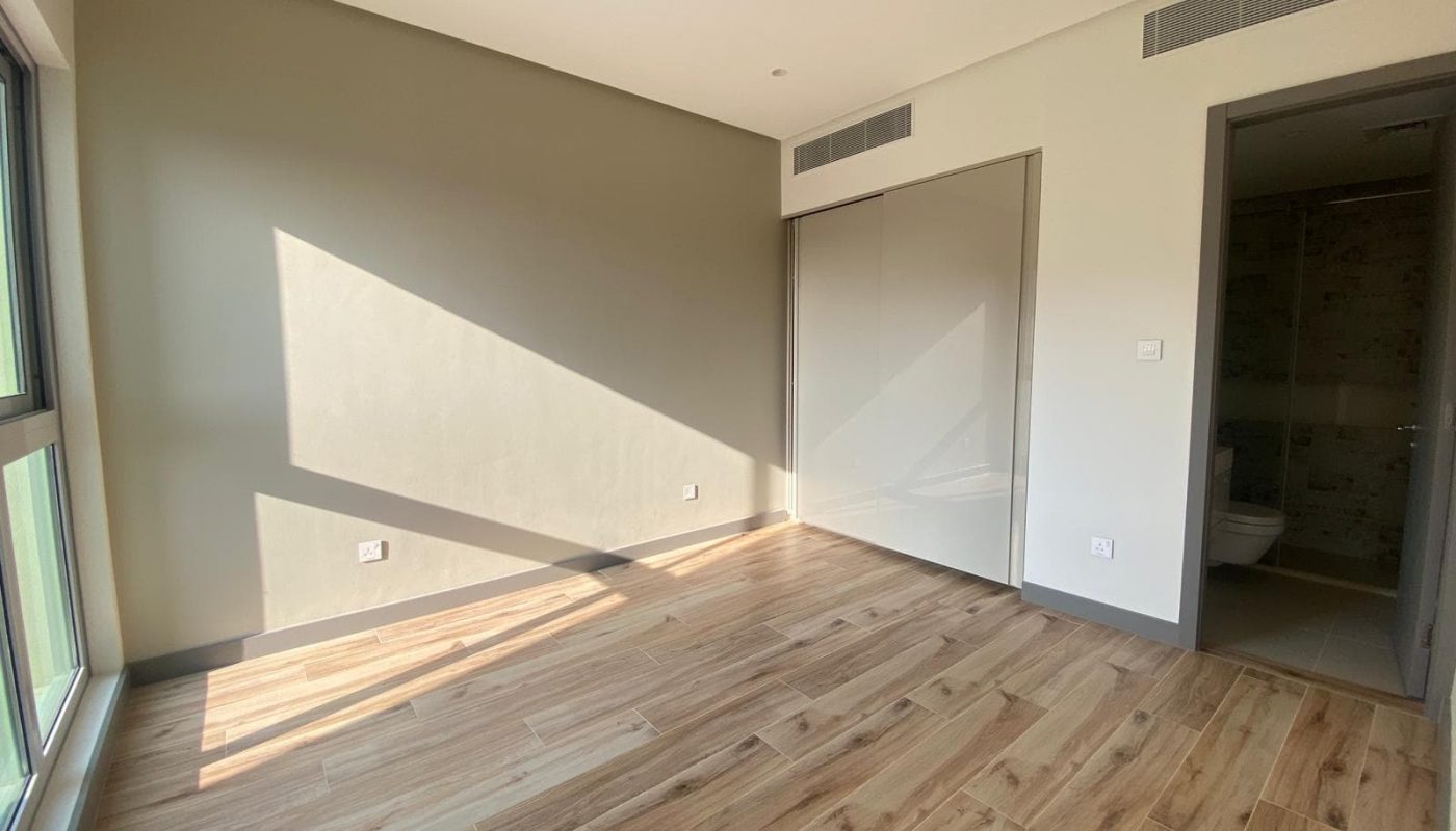 Empty room with wooden flooring, large window on the left, sunlight casting shadows on the wall, built-in closet with sliding doors, and an open door leading to a bathroom.