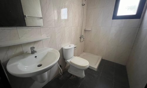 A minimalist bathroom with a sink, toilet, and shower area. The walls are tiled in a light color, and the floor features dark tiles. A small window provides natural light.