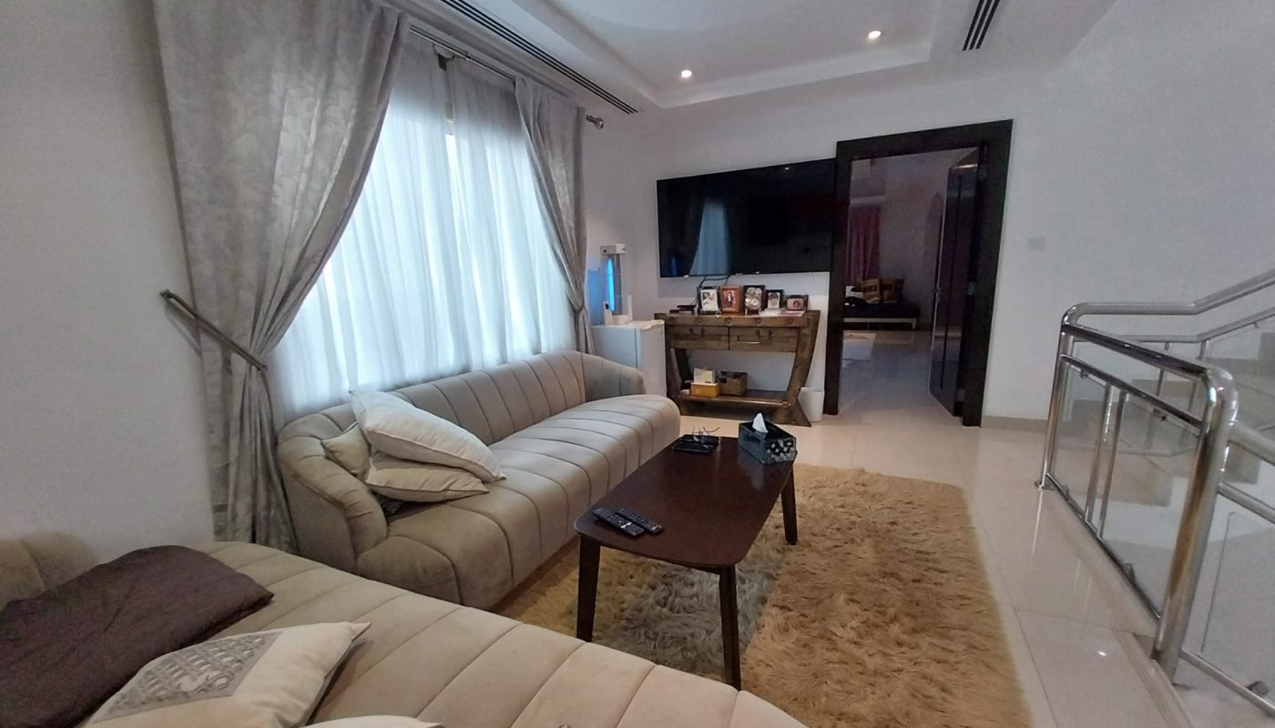 Modern living room with beige sofas, a wooden coffee table, a shag rug, and a TV on the wall. Large window with curtains lets in natural light. A staircase and hallway are visible.