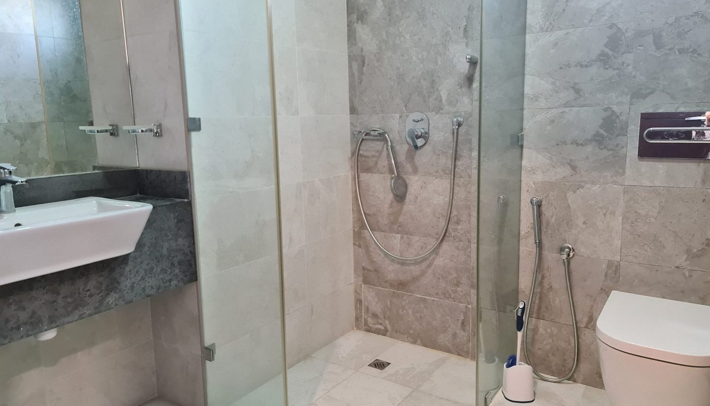 Modern bathroom with a glass-enclosed shower, wall-mounted toilet, sink with countertop, and gray stone tiles. Showerhead and faucet are visible. Toilet brush and spray hose present.