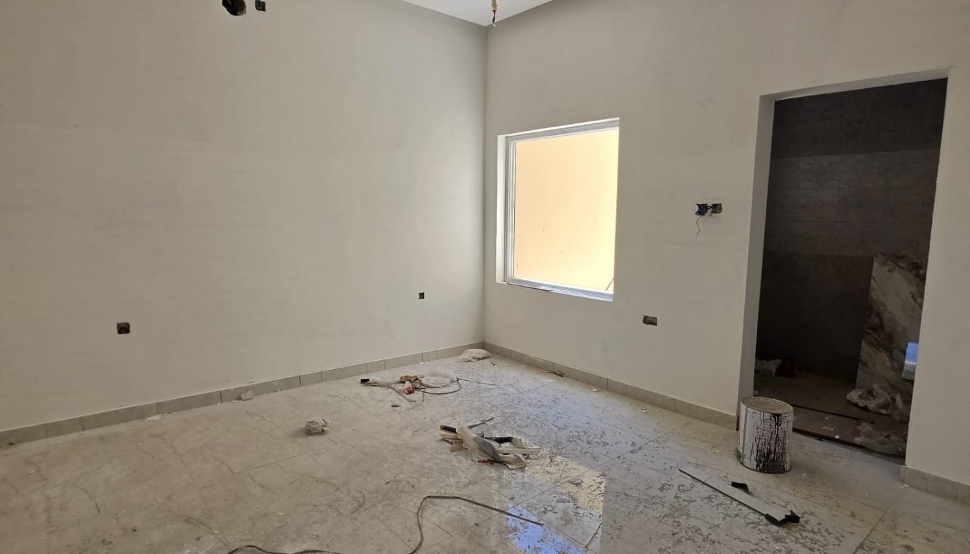 Unfinished room with tiled floor, exposed wires, construction materials, and an open doorway leading to another area, with sunlight coming through a single window.