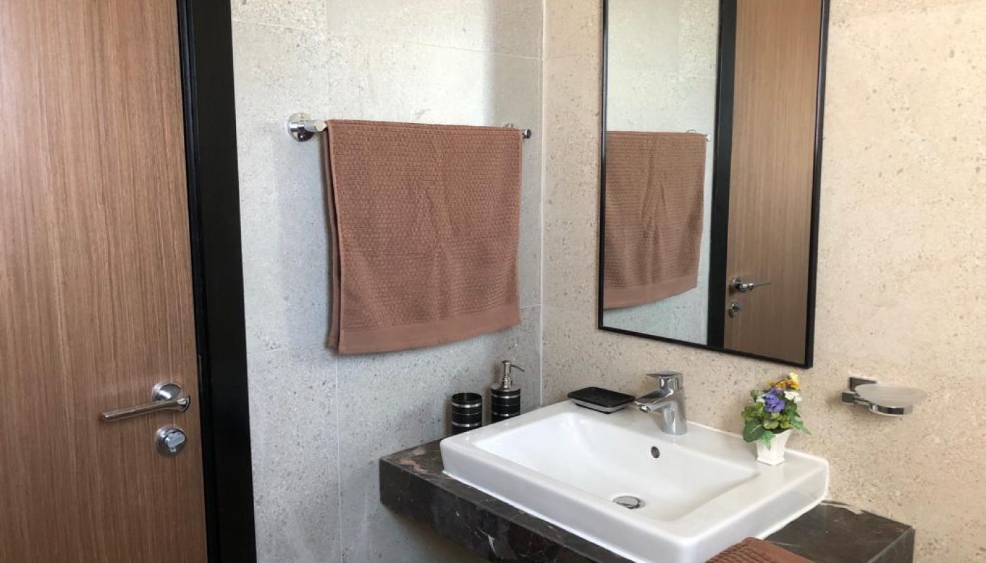 Modern bathroom with a rectangular mirror, brown towel on a rail, white sink on a dark countertop, soap dispenser, and a small vase with flowers.