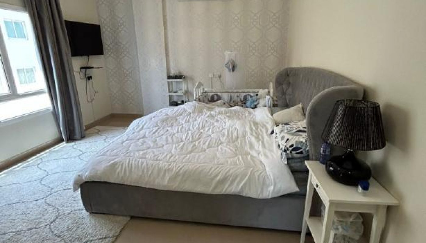 A bedroom with a large bed, nightstand, lamp, wall-mounted TV, air conditioner, and a window with curtains, featuring light-colored walls and tiled floors.