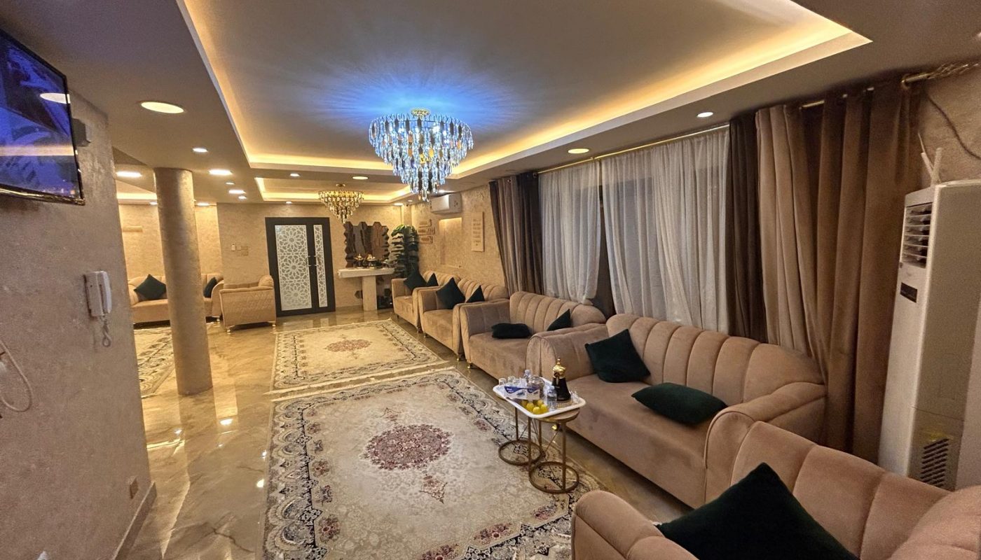 A well-lit living room with beige sofas, a chandelier, patterned rugs, a TV on the wall, and a coffee table with drinks and snacks. Large curtains cover the windows.