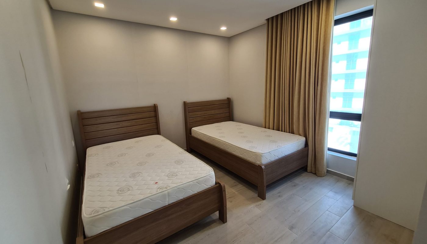 Room with two wooden single beds, mattresses, beige curtains, and a large window. Walls and floor are light-colored. Ceiling lights illuminate the space.