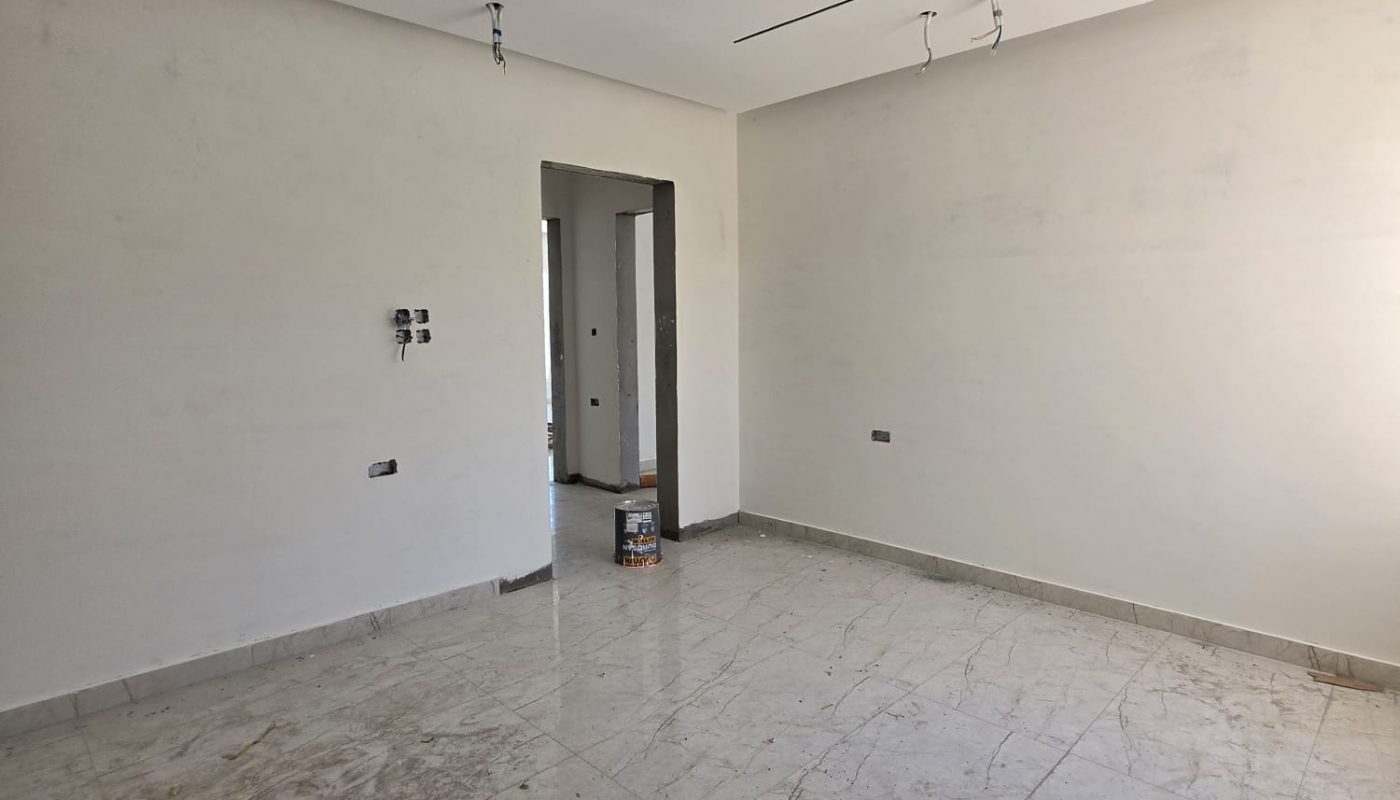 Empty, unfurnished room with tiled floor, white walls, exposed ceiling wires, and a small box placed near the doorway to another room.