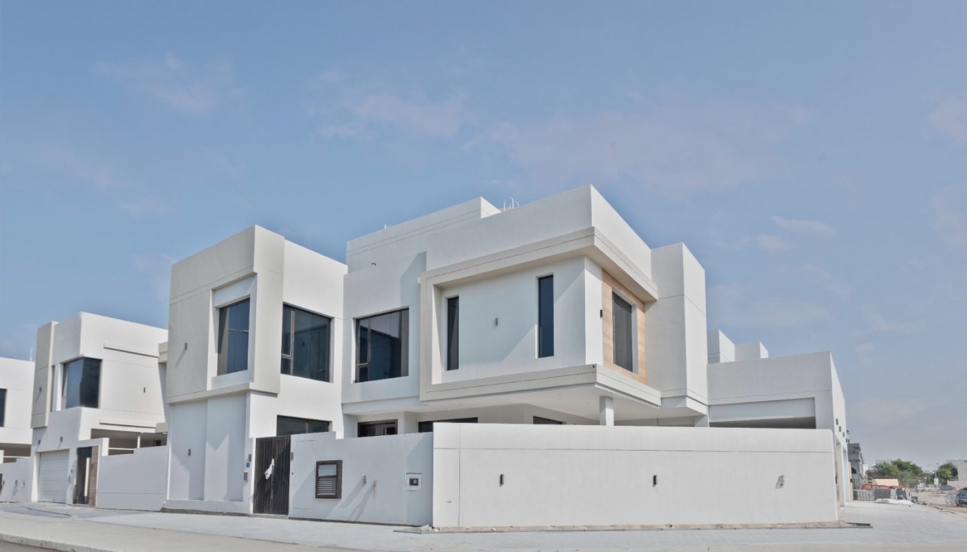 Modern white two-story house with large windows and flat roof, situated on a corner lot with a surrounding wall, under a clear blue sky.