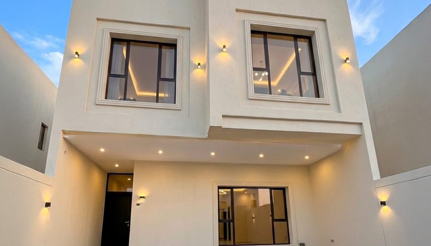 Modern two-story beige house with large windows, exterior wall lights, and a spacious tiled front yard under a clear blue sky.