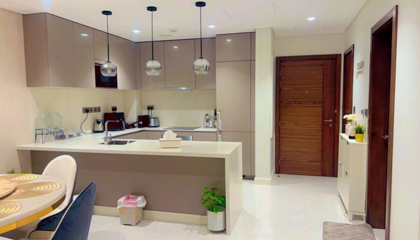Modern kitchen with beige cabinets, white countertops, and stainless steel appliances. Three pendant lights hang above the island with a sink. Dining table with chairs nearby.