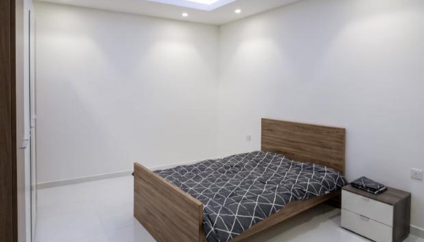 Minimalist bedroom with a wooden bed, geometric-patterned bedding, a nightstand with items, white walls, tiled floor, and recessed ceiling lights.