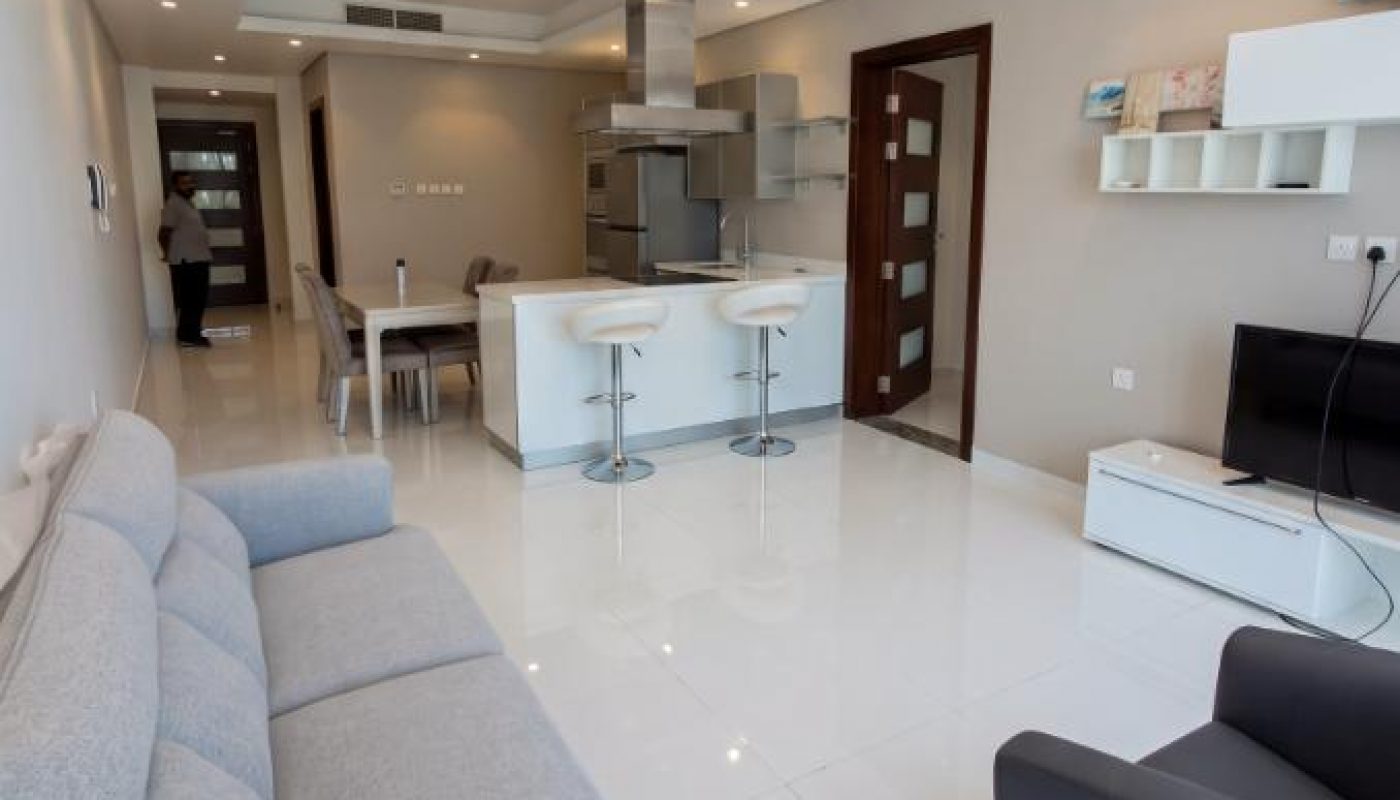 Modern open-plan living room and kitchen with white floors, gray furniture, bar stools, TV, and wall shelves. A person stands near the dining table in the background.