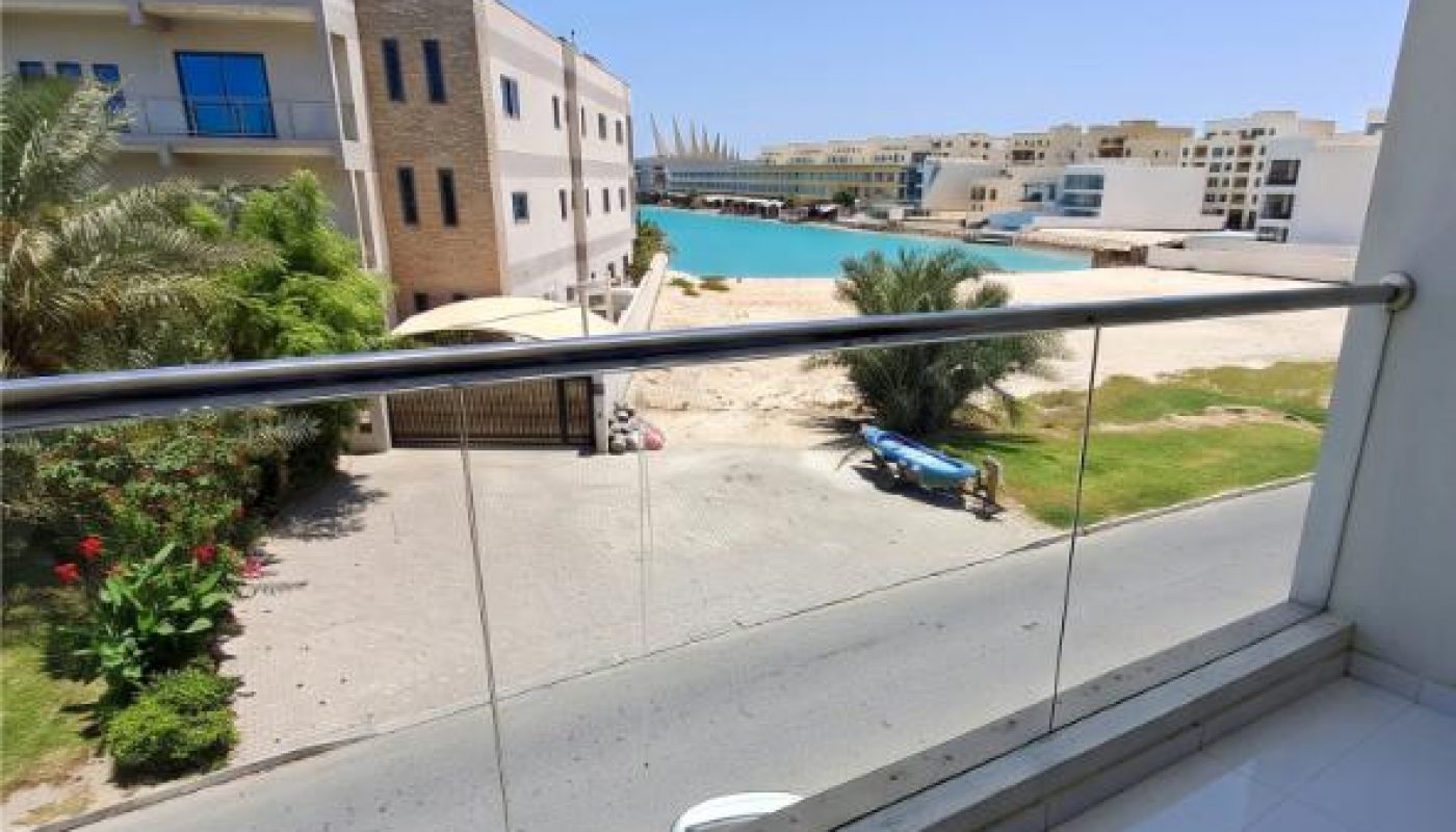 View from a glass balcony overlooking a road, garden, and waterfront with modern buildings under a clear blue sky.