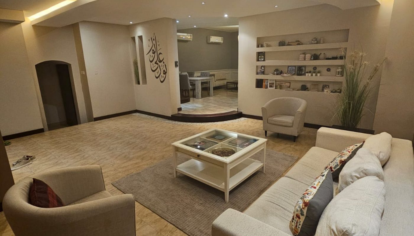 Modern living room with beige sofas, a glass coffee table, built-in shelves, and soft ceiling lighting, leading to an open dining area in the background.